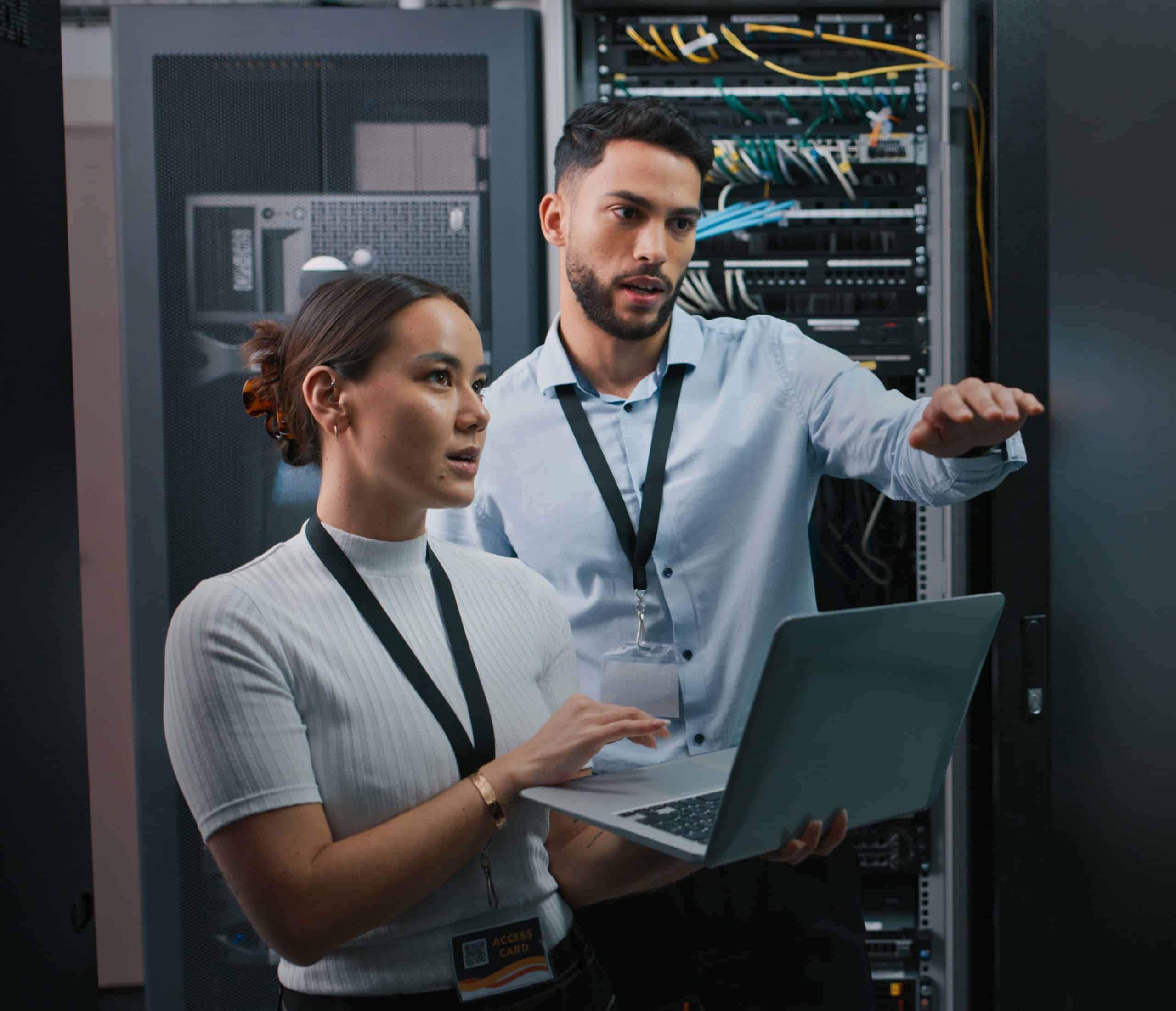 Two IT professionals in a data center review server equipment, with the man pointing at cables while the woman holds a laptop