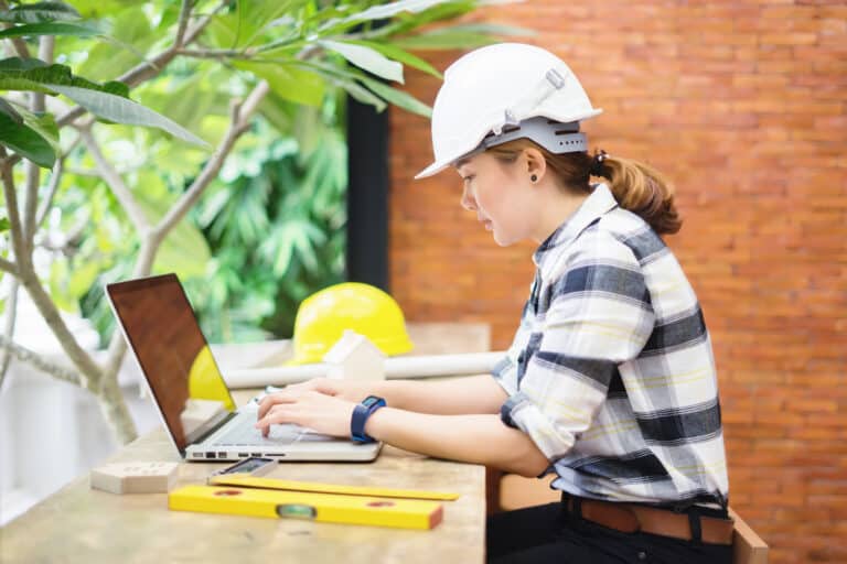 A female construction worker sits at a laptop outdoors.
