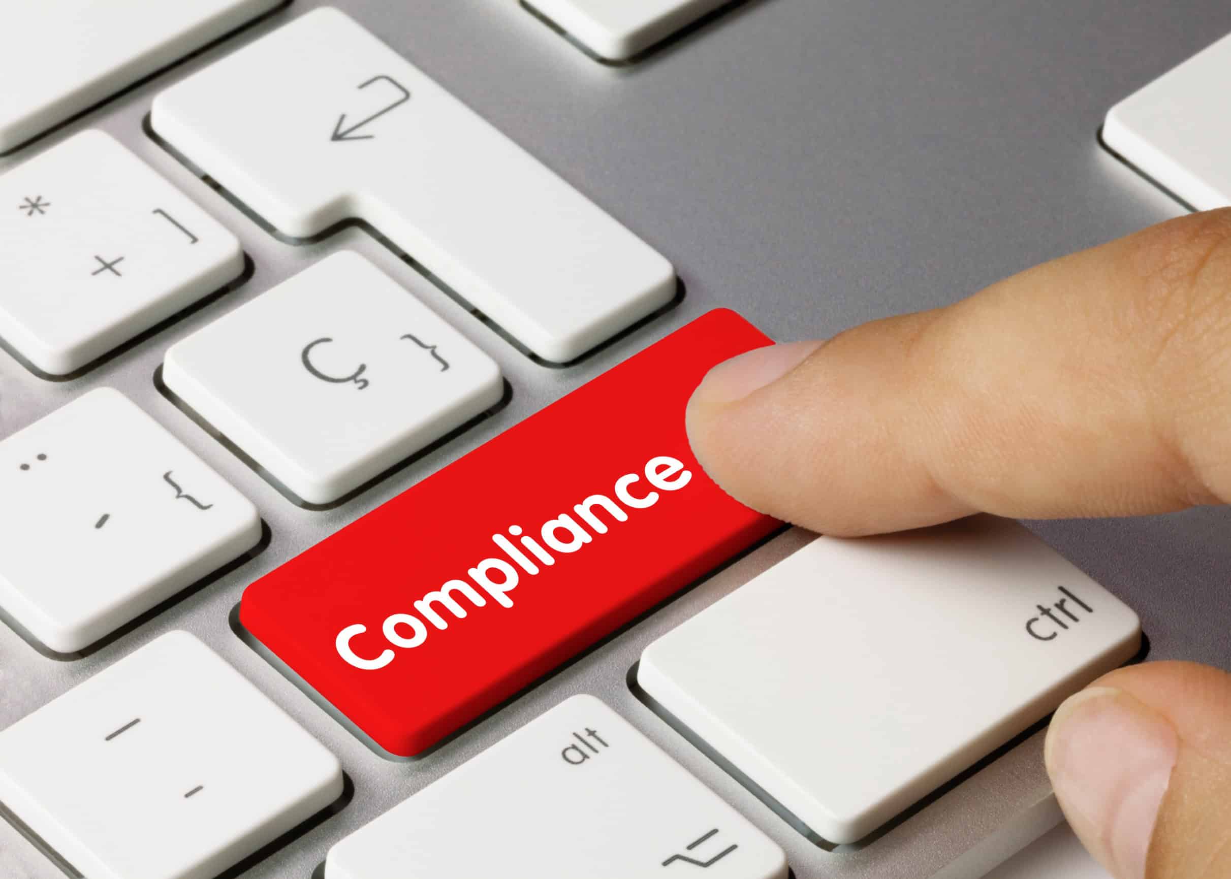 A finger pushes a red button on a white keyboard that says “Compliance.”