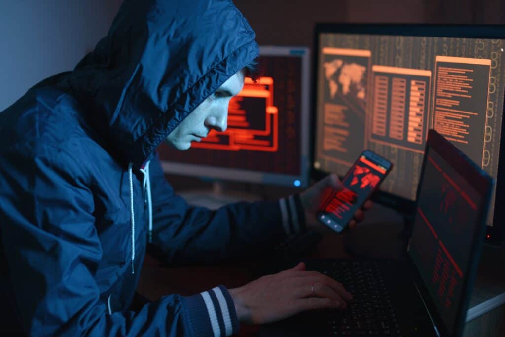 A male hacker in a hoodie using a computer in the dark under neon light, holding a phone. Concept of cyberattack.