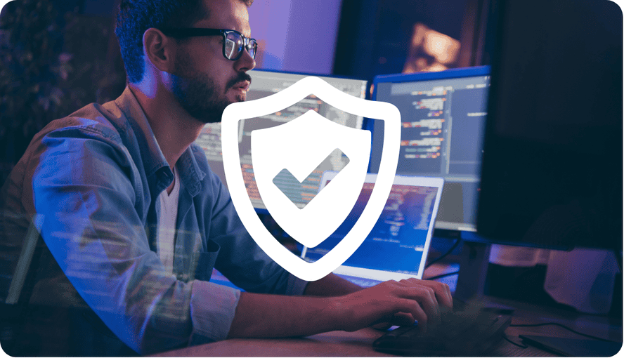 A man in glasses at a multi-monitor computer setup with a white security shield and checkmark overlay, symbolizing cybersecurity.