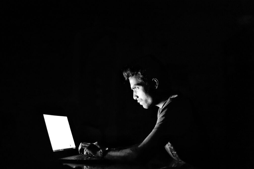 Man seated at a desk in a black room, hunched over a computer with a very bright screen.