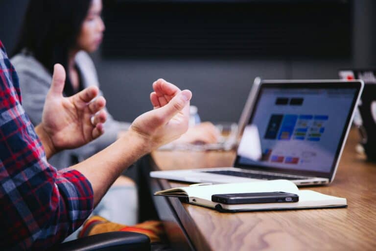 A view of someone's hands gesturing while they are talking to coworkers seated at a conference table with a laptop in front of them.