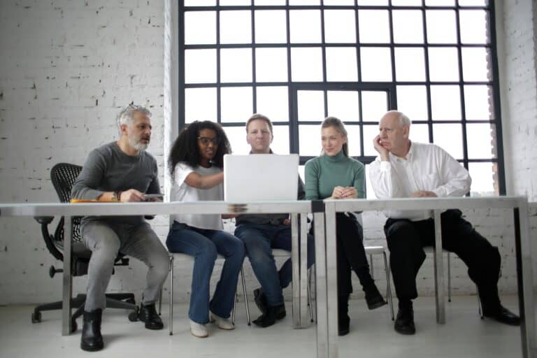 Five business professionals sitting next to eachother at a table in an office, looking and pointing at a laptop screen.