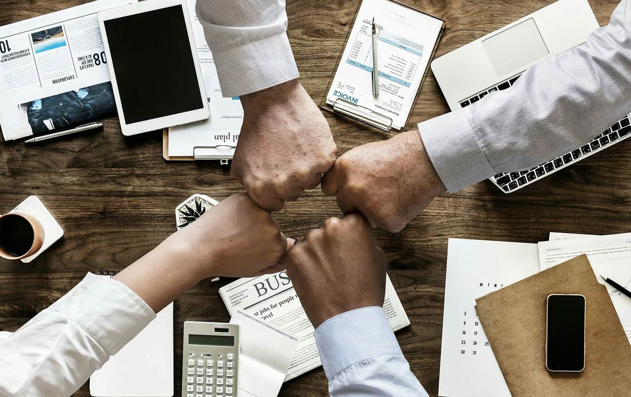 Four workers arms reaching out for a fist bump over a desk with scattered paperwork and digital devices.