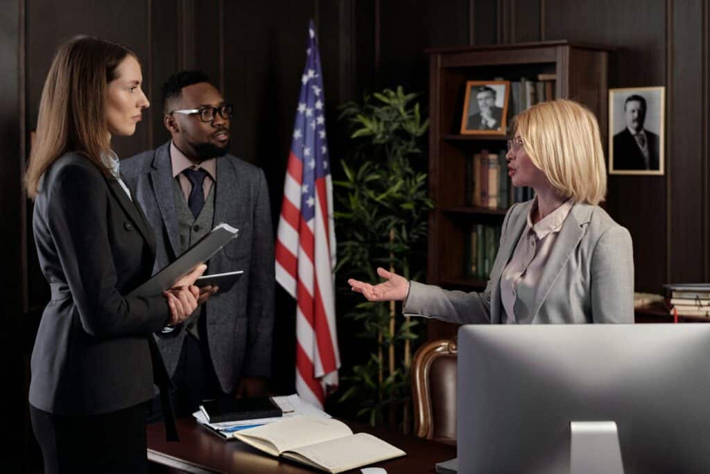 Three law firm employees amidst conversation, standing in an office with an American flag and a large desktop computer