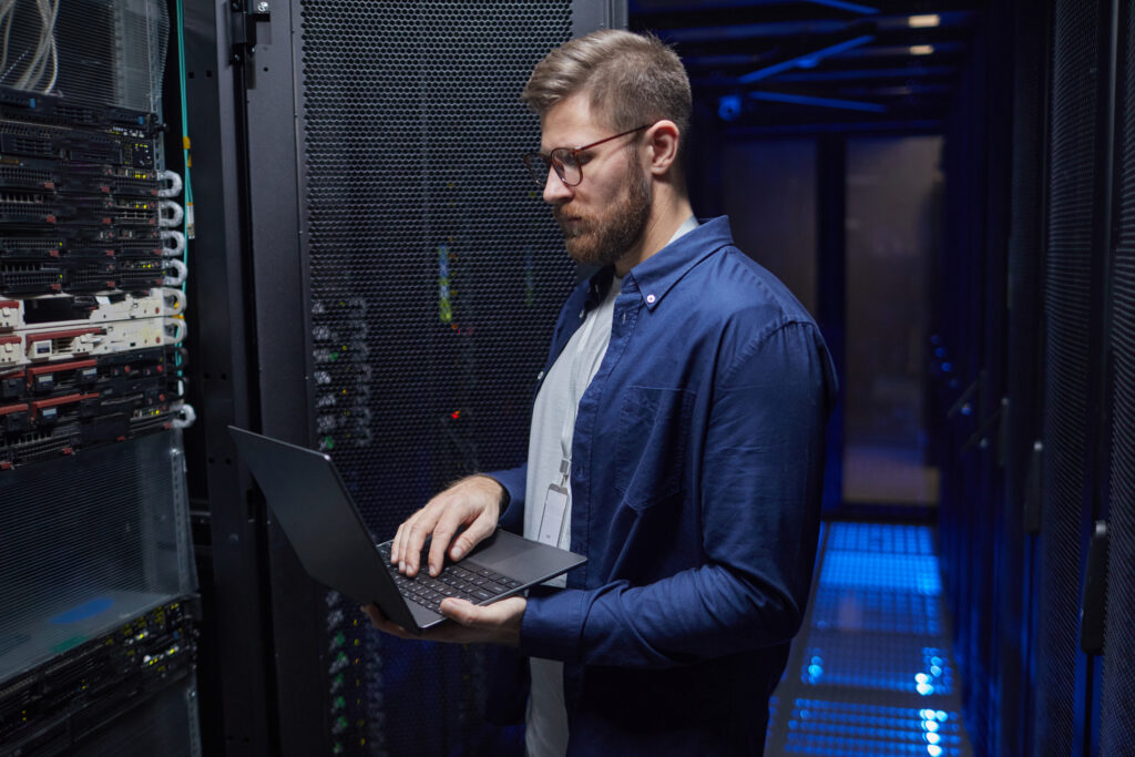IT technician wearing glasses and a blue shirt uses a laptop in a data center beside server racks.