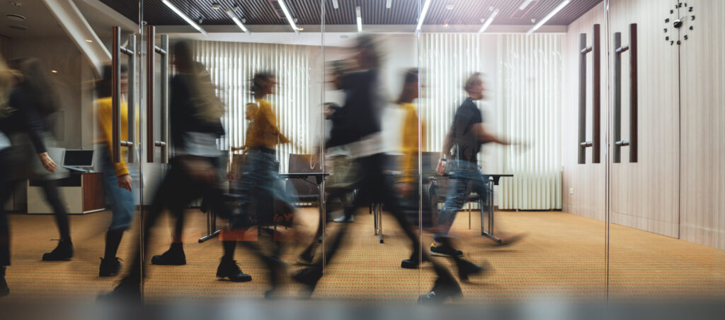 Busy office scene with motion-blurred coworkers walking past glass-walled meeting rooms and vertical blinds in a modern space.