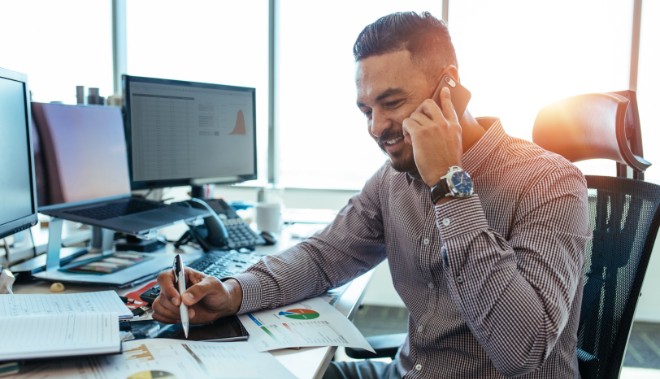 Smiling man in a checkered shirt sits at a desk, talking on his phone while taking notes on charted documents.