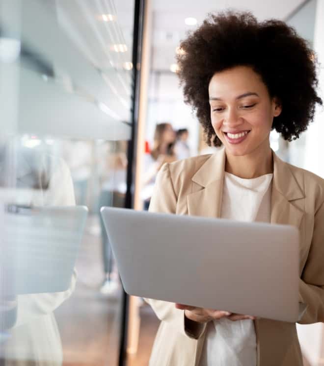 Smiling woman in beige blazer holds a laptop in a bright office hallway, looking at the screen.