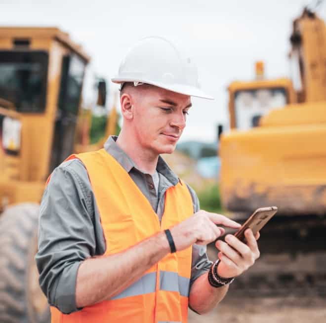Construction worker in an orange safety vest and white hard hat using a smartphone at a construction site with machinery in the background.
