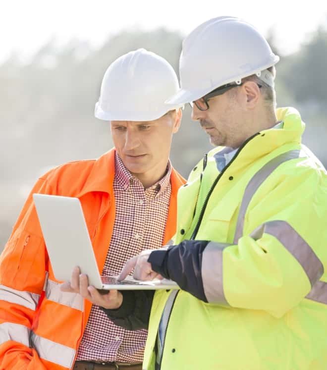 Two construction workers wearing hard hats and high-vis jackets review data on a tablet at a worksite.