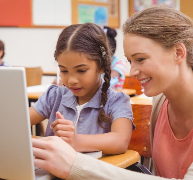 Teacher helps a young girl use a laptop in a bright classroom.