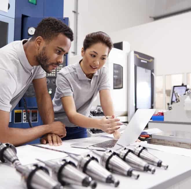 Two technicians in a factory lean over a laptop, reviewing data while inspecting metal tooling on a workbench.