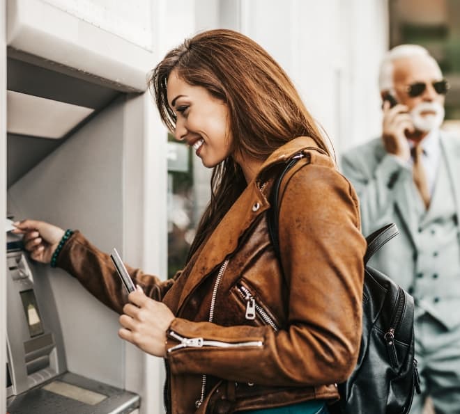 Woman in a brown leather jacket uses an outdoor ATM, holding her card with a backpack on her shoulder, smiling; a man in a suit talks on his phone in the background.
