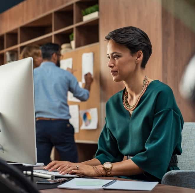 Professional woman typing at a desk computer in an office; coworker pins notes on a corkboard behind her.
