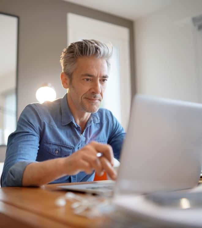 Man with gray hair in a denim shirt working on a laptop at a wooden desk in a bright room, concentrating on his screen.