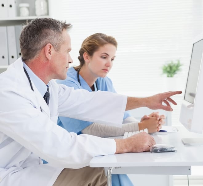 Two healthcare professionals, a doctor and a nurse, review information on a desktop computer together in a bright clinic.