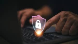 A remote worker touches a shield with a lock over their laptop keyboard that depicts cybersecurity.