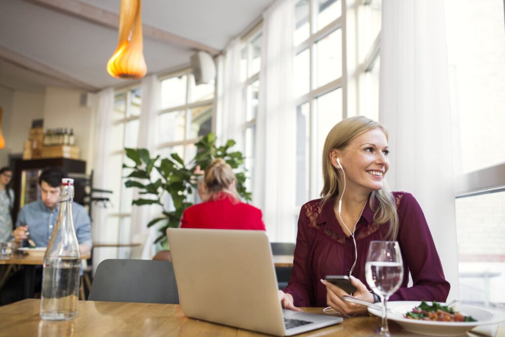 Woman in café smiling while working on laptop with phone and earphones