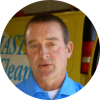 Man in a blue shirt speaking in front of a yellow vehicle or storefront. Colors and setting suggest a casual interview or press appearance.
