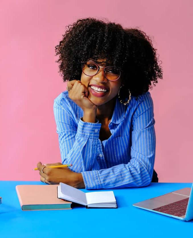 Smiling woman with curly hair and glasses sits at a blue table with a laptop, notebook, and pencil against a pink background.