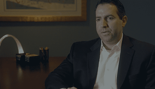 Man in a dark suit sits at a desk in an office, looking ahead with a neutral expression.