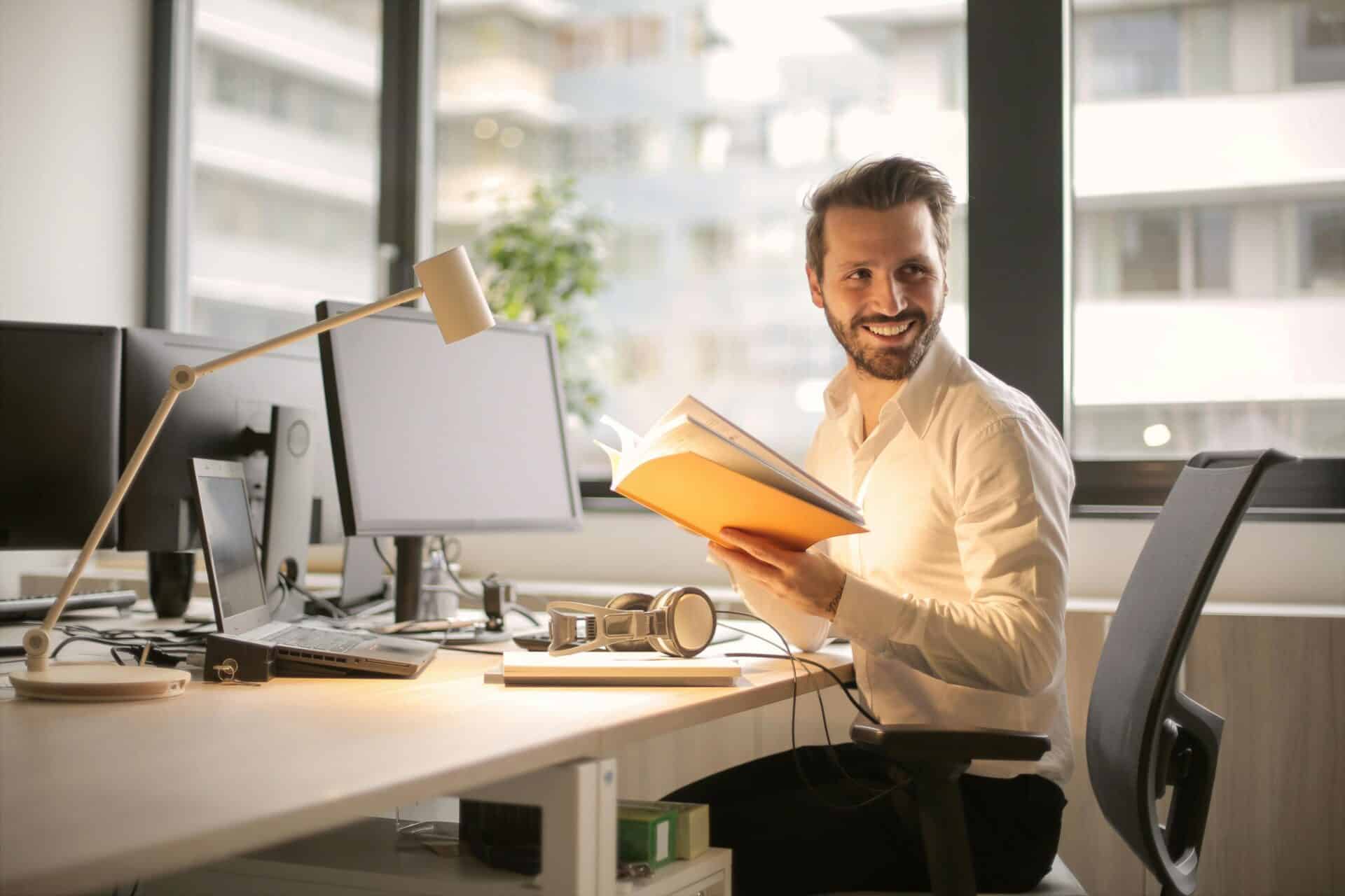 A male cybersecurity expert from a Security Operations Center sits at a desk in front of a monitor and holds a notebook in his hands.