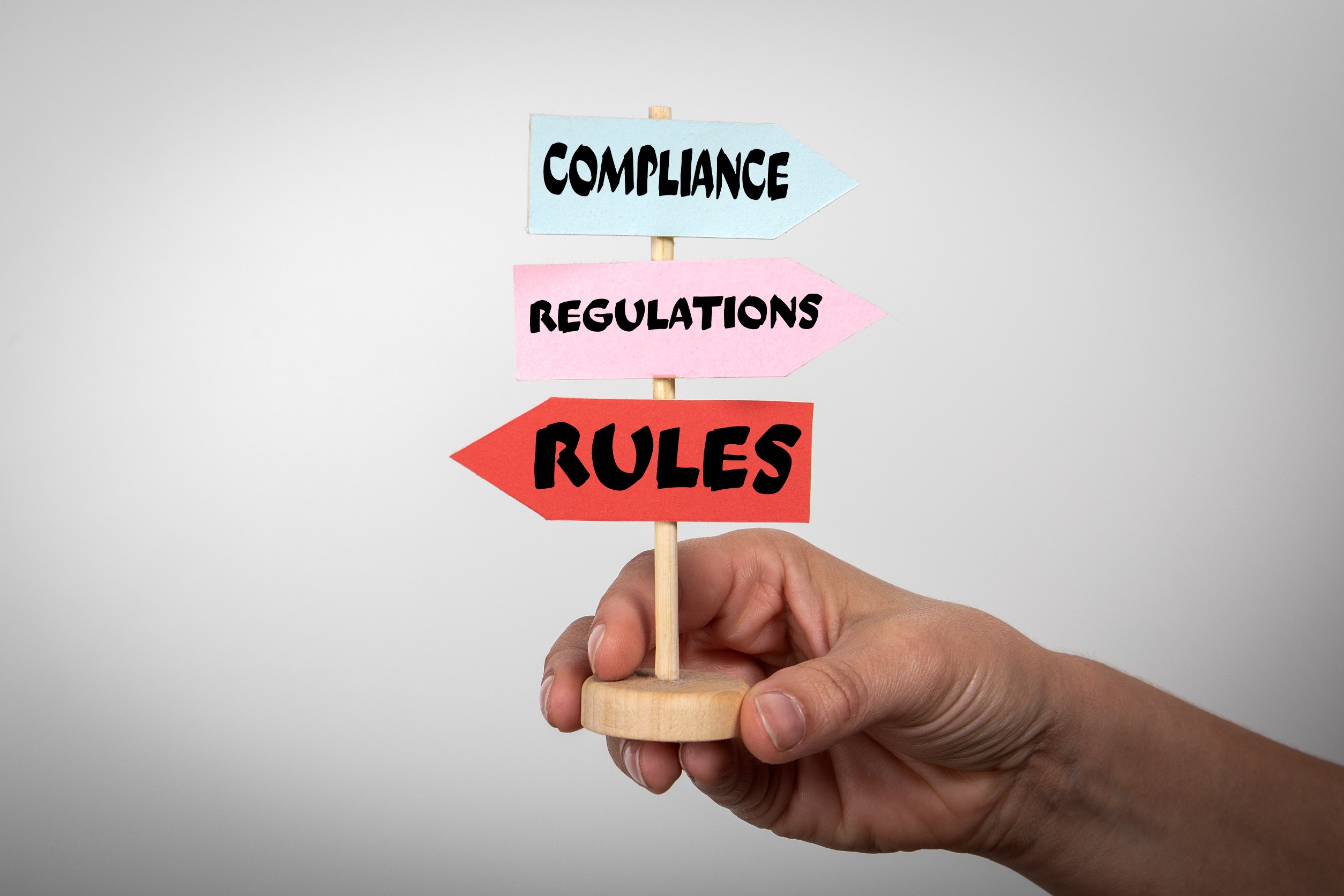 A hand holding a small wooden signpost with arrows labeled "Compliance," "Regulations," and "Rules."