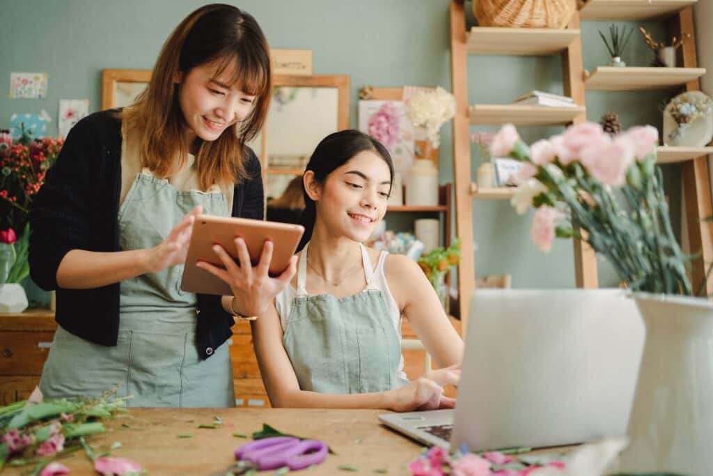 A business owner works at a computer and holds up a tablet to show their employee some information