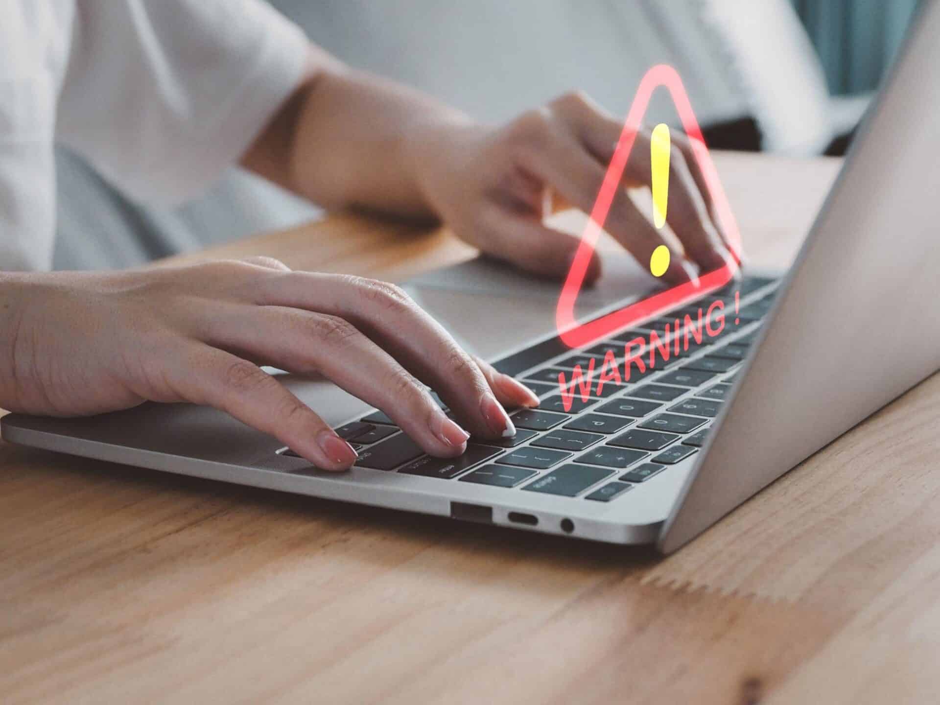 Two hands type on a laptop keyboard as a holographic image of a warning appears above.