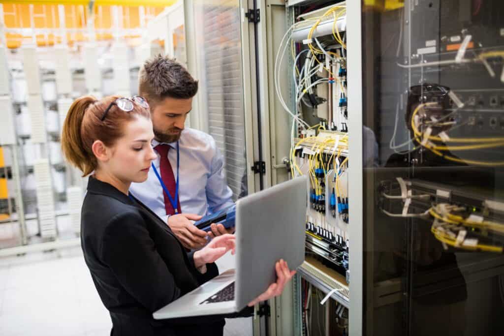 Two network management professionals in business attire focused on a laptop screen in the server room.