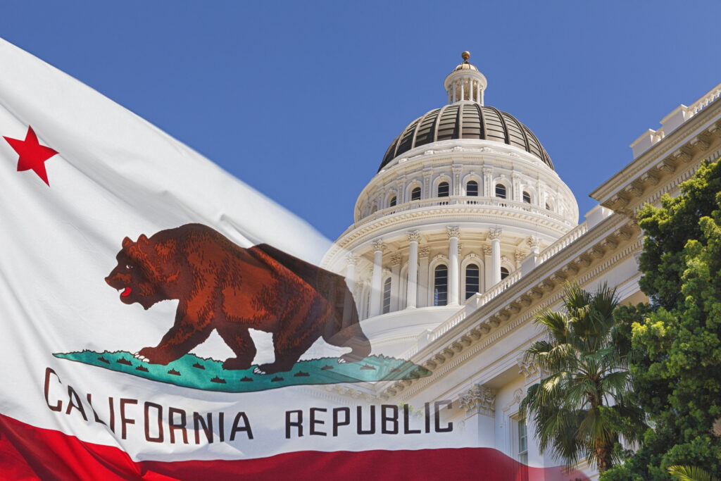 California state flag waving in the foreground with a brown bear image and red stripe, with the California Capitol dome and blue sky behind it.