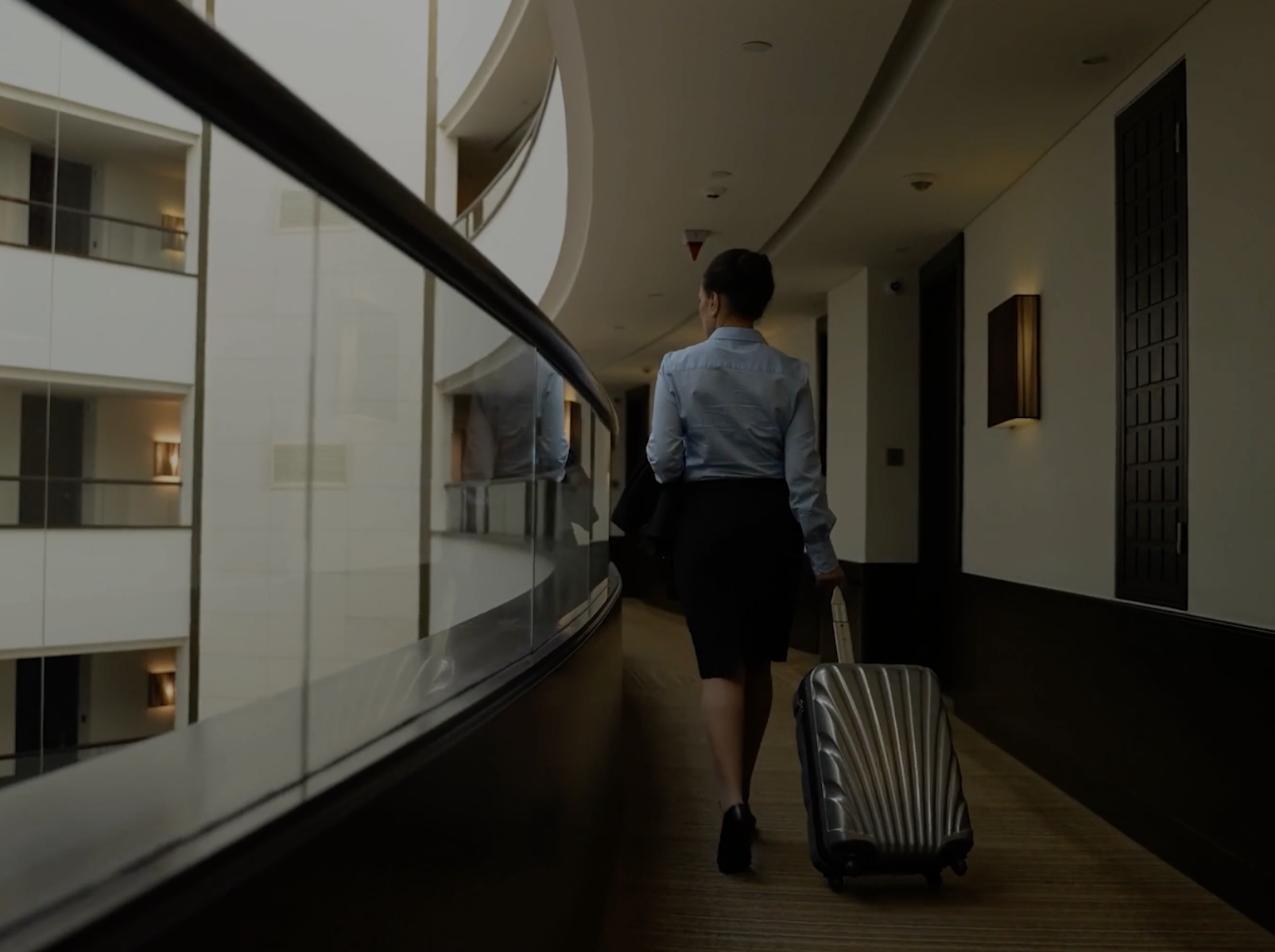 Woman walking down a curved hotel corridor, pulling a silver rolling suitcase behind her (back view).