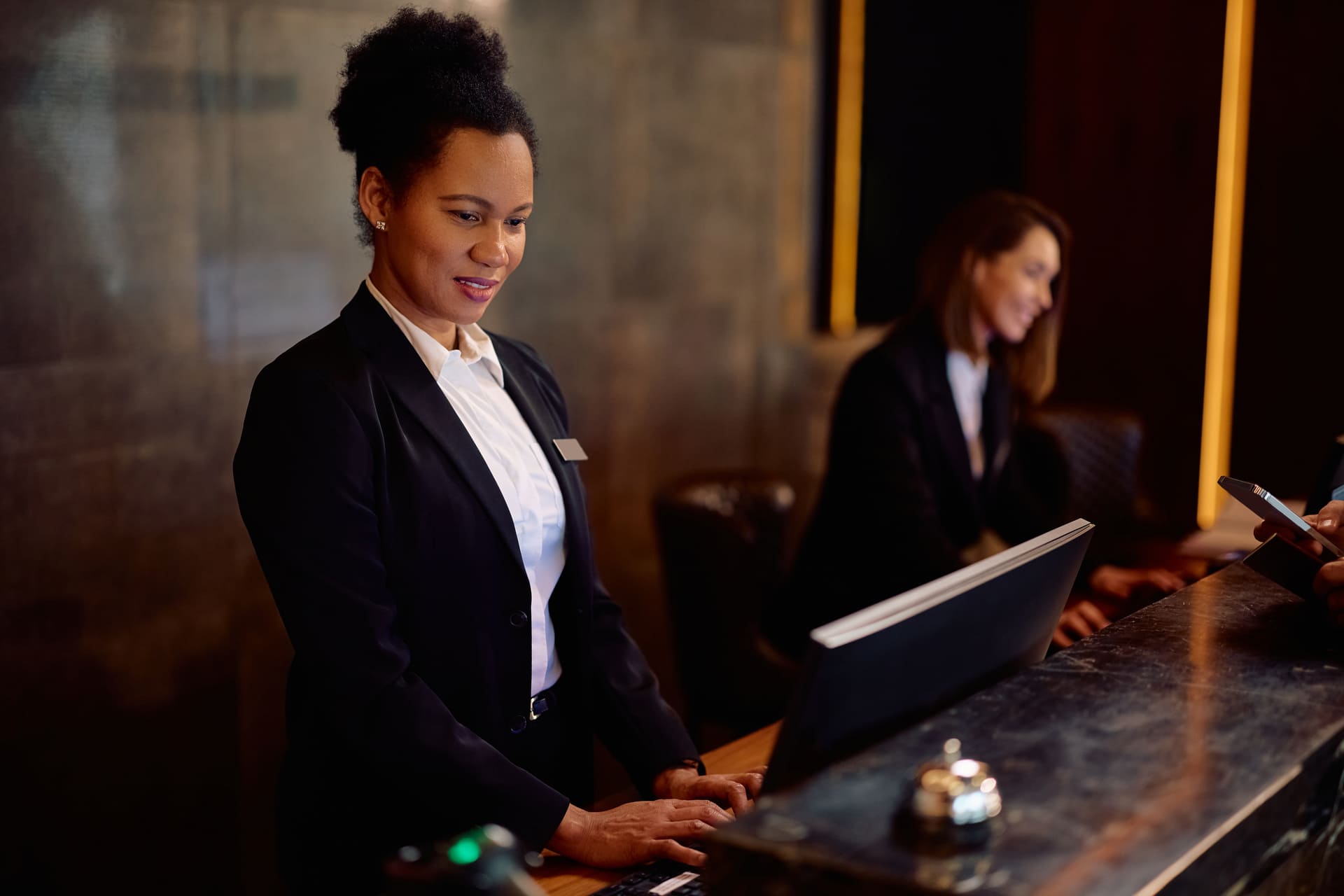 Front desk receptionist in a black blazer and white shirt at a marble counter, looking at documents; another staff member in the background.