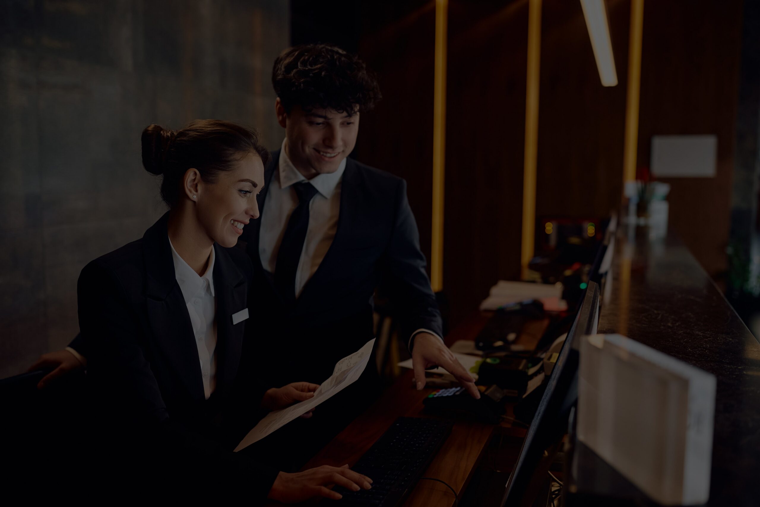 Two receptionists in dark suits work at a front desk, reviewing documents on a laptop in a dim, modern lobby setting.