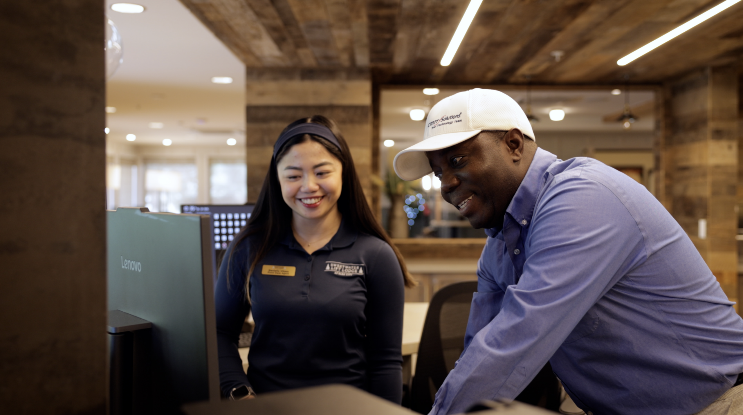 Two coworkers lean over a computer monitor, smiling as they review the screen (woman in navy polo, man in a white cap and blue shirt).