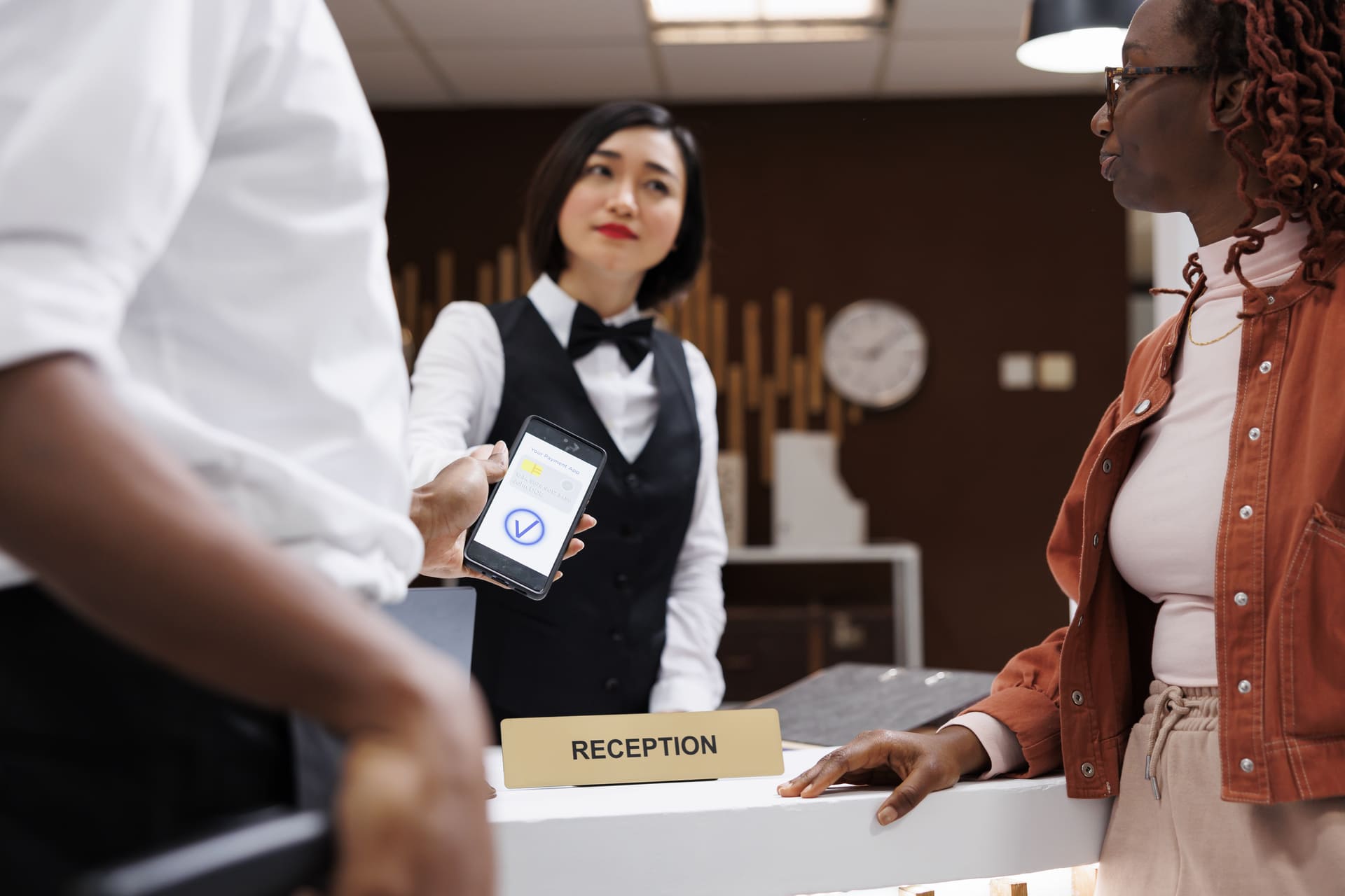 Reception counter scene with a uniformed attendant showing a smartphone to a customer for payment or verification