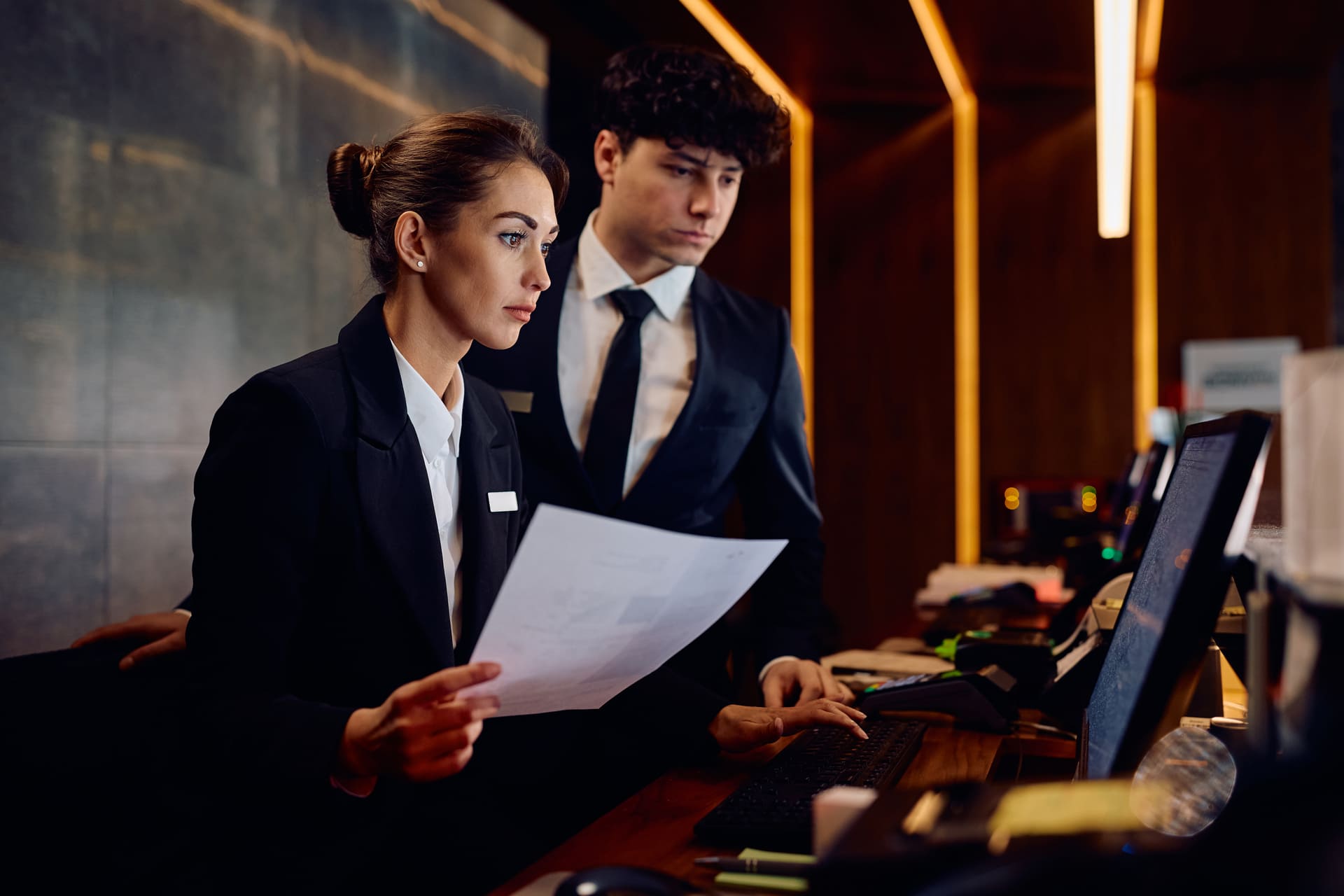Two hotel receptionists in business attire stand at a front desk, one holding a document while the other types on a computer.