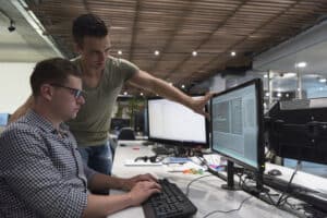 Two male coworkers perform a security audit together on a dual-monitored computer.