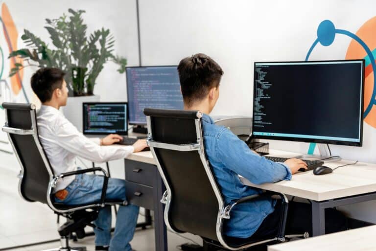 Two men sit at desks engaging with their computers in a collaborative workspace.