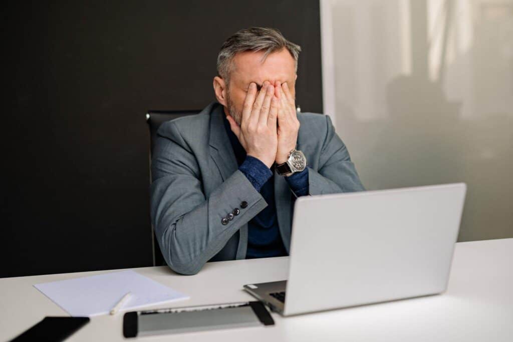 A businessman sitting in front of a laptop covers his face in frustration.
