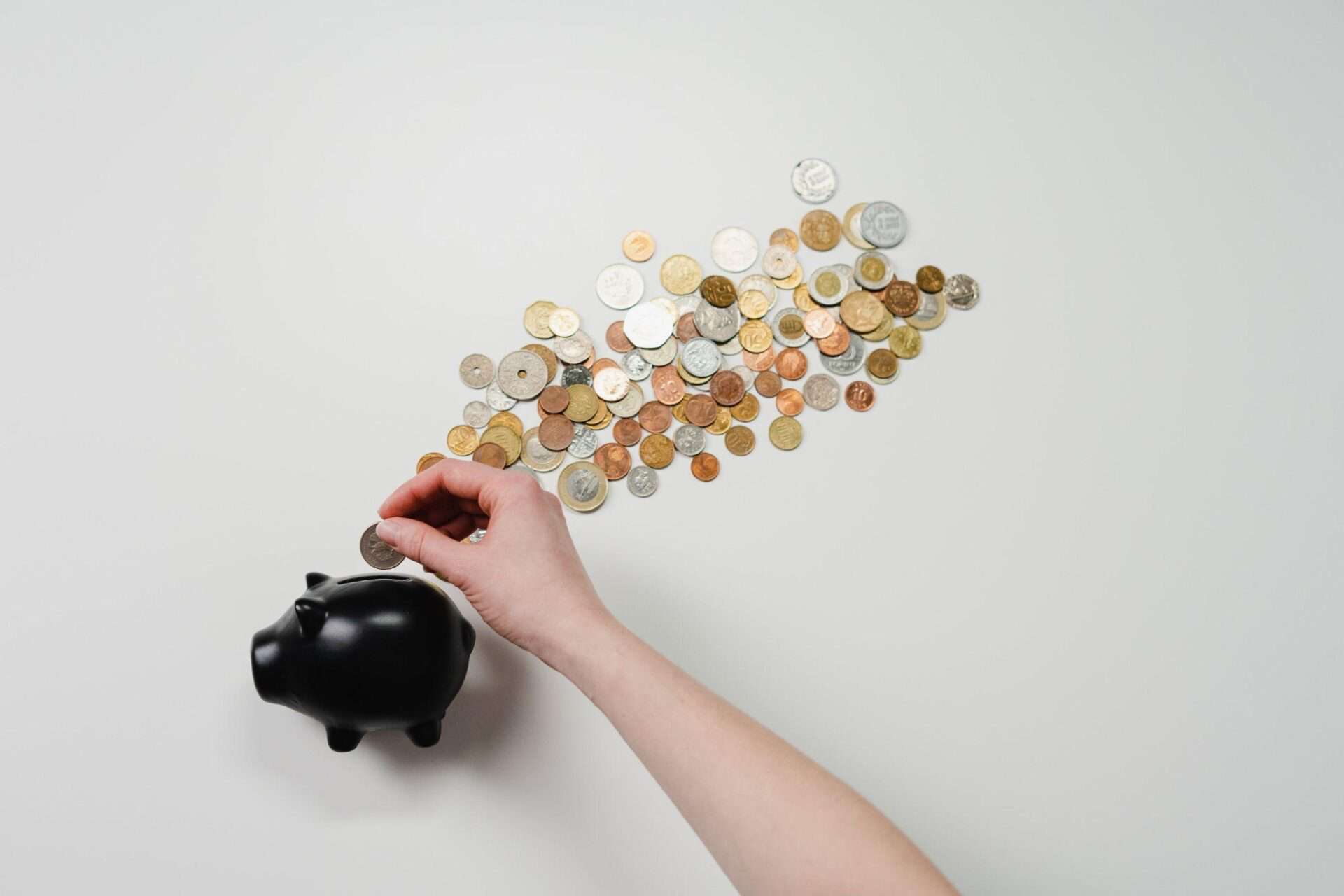 A hand puts a coin into a black piggy bank as coins spill out of it.