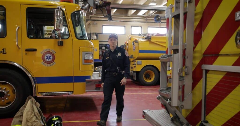 Firefighter in uniform stands between yellow fire trucks inside a fire station bay with a patch on the door of the nearest truck.