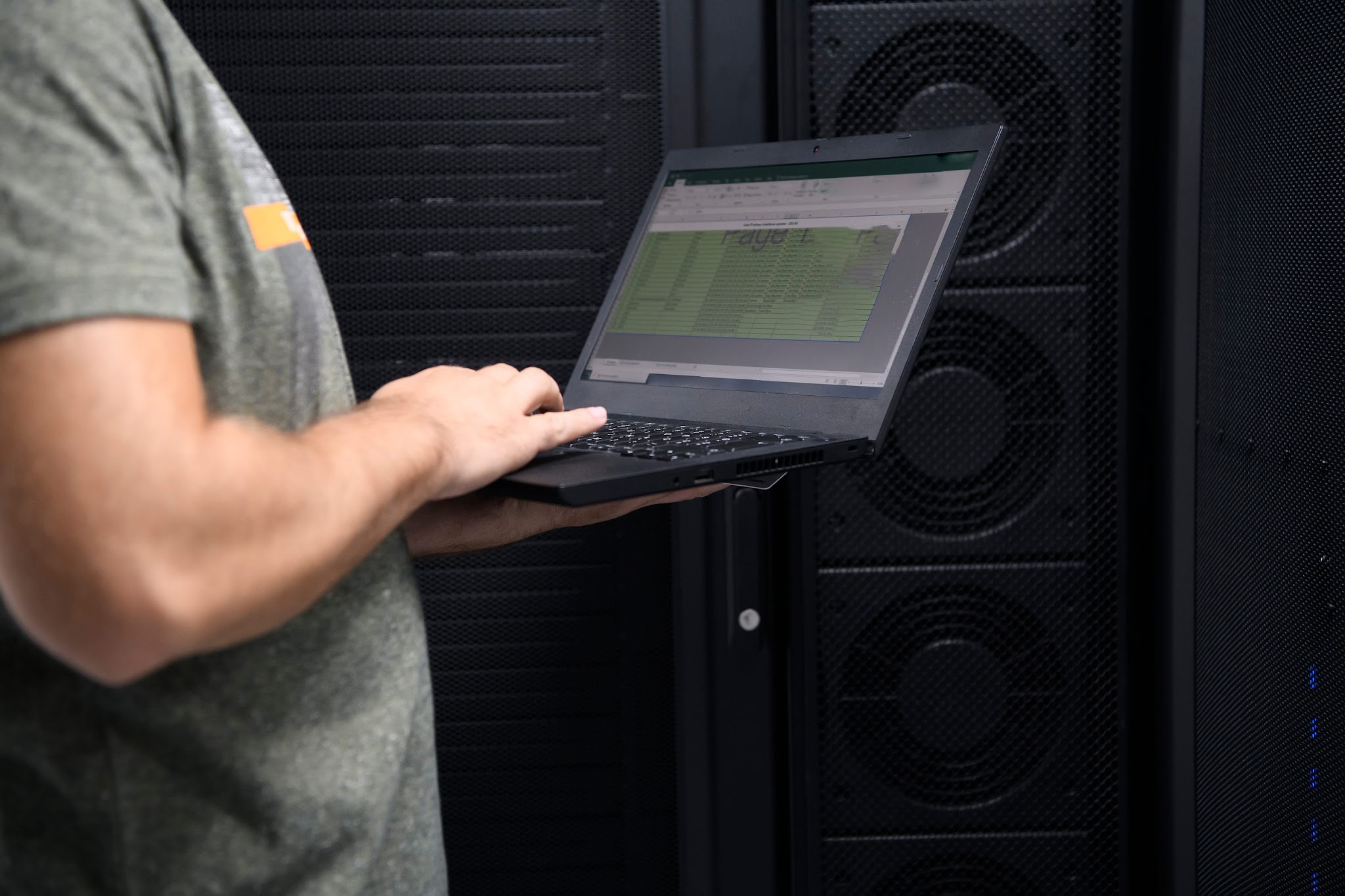 IT technician standing in a server room holding a laptop displaying a network monitoring spreadsheet, representing hands-on proactive IT management for North Pittsburgh businesses.