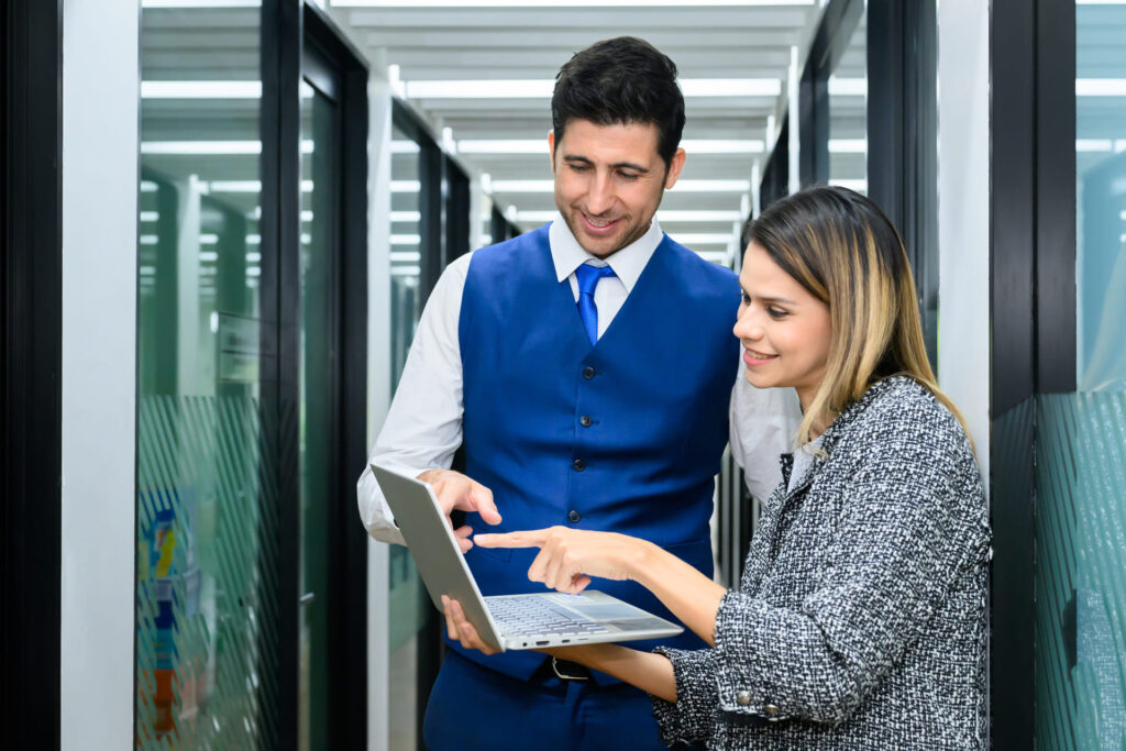 Two business professionals discussing work on a laptop in office hallway