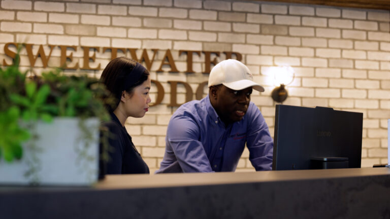 Two coworkers review something on a computer at a reception desk against a brick wall backdrop.