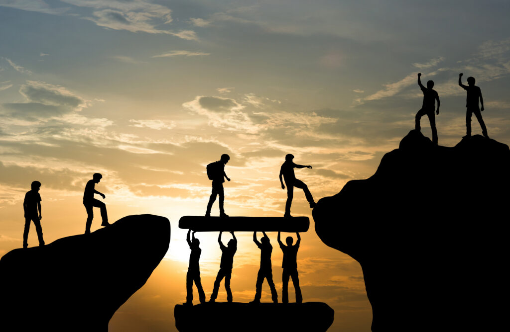Silhouettes of people helping each other across a rocky chasm at sunset, reflected in the water below.
