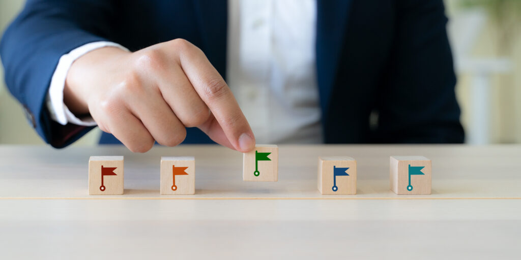 Businessperson in a suit places a wooden block with a green flag among a row of flagged blocks on a table, symbolizing stepwise progress.