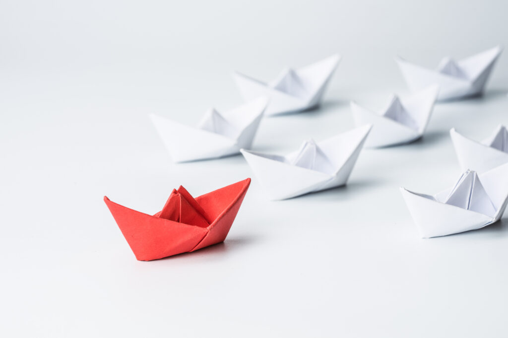 Red origami boat in the foreground with several white origami boats blurred in the background on a white surface.